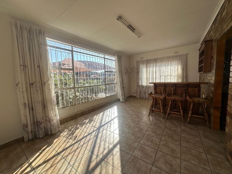 A living area featuring a built-in wooden bar, tiled flooring, and large windows with security bars, offering a functional space for entertainment.