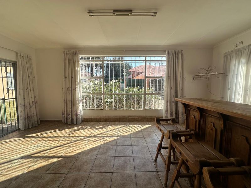 A living area featuring tiled flooring, a built-in wooden bar counter, and large windows with security bars overlooking the garden.