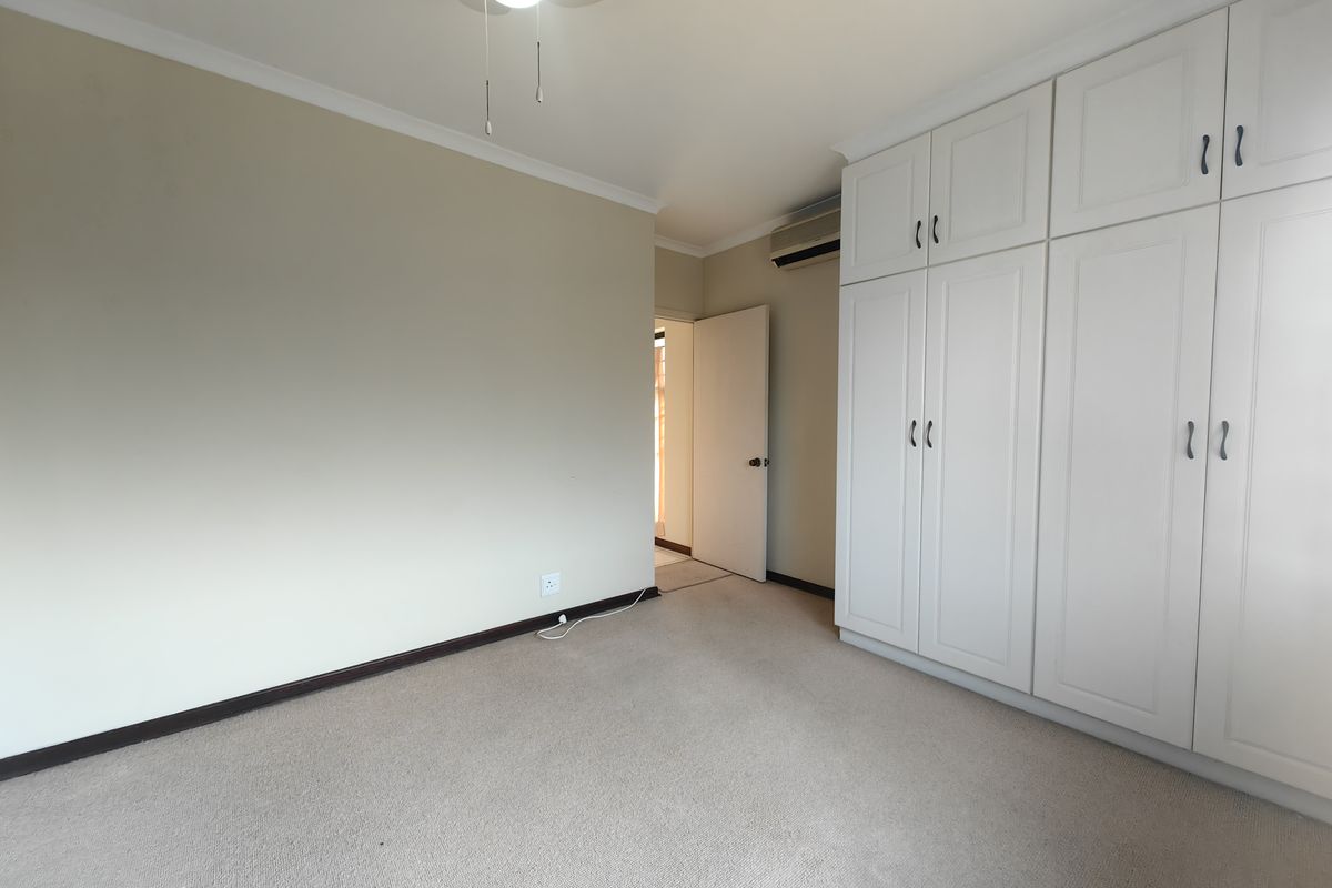 A clean, empty bedroom featuring neutral carpeting, ample built-in cupboard storage, a ceiling fan, and a wall-mounted air conditioning unit.