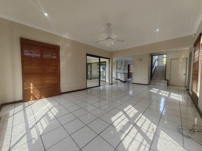 Spacious open-plan living area featuring neutral tiled flooring, a ceiling fan, and direct access to the kitchen and staircase.