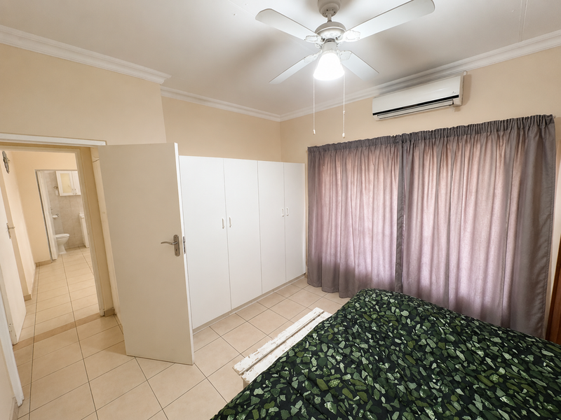 A bedroom featuring built-in cupboards, a ceiling fan, and a wall-mounted air conditioning unit, with direct access to an adjacent bathroom.