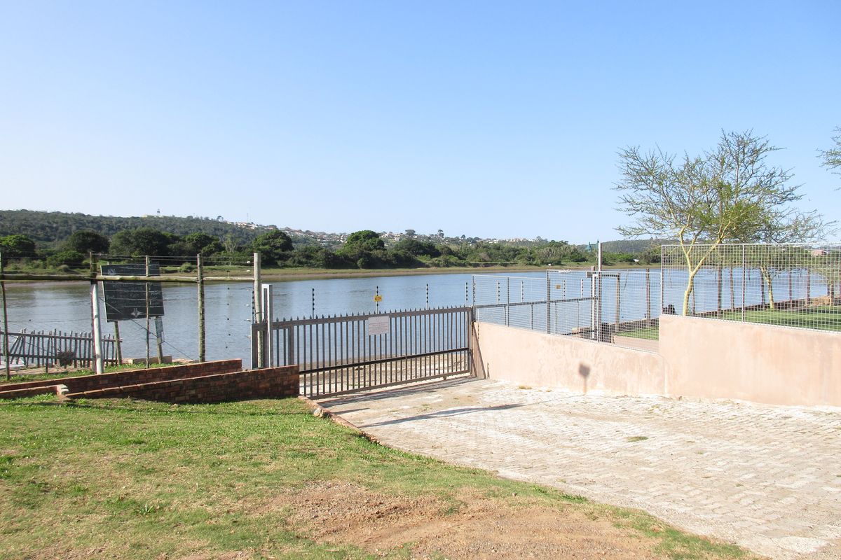 A view of the rear property boundary featuring a paved driveway, metal security gate, and electric fencing overlooking a river or lagoon.