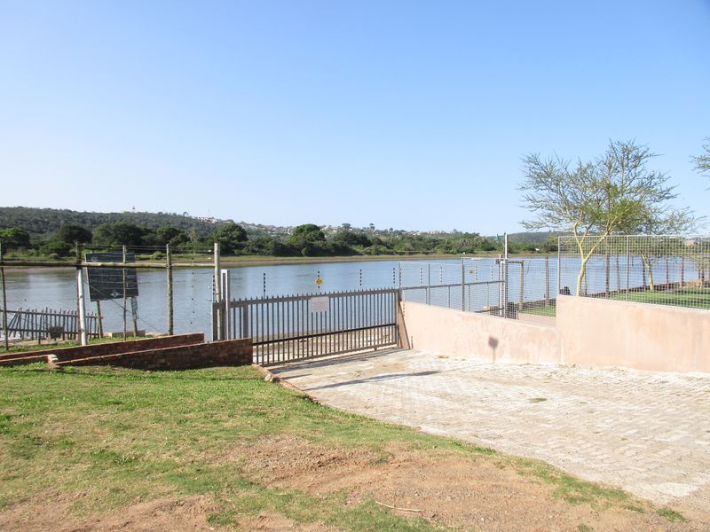 A view of the rear property boundary featuring a paved driveway, metal security gate, and electric fencing overlooking a river or lagoon.