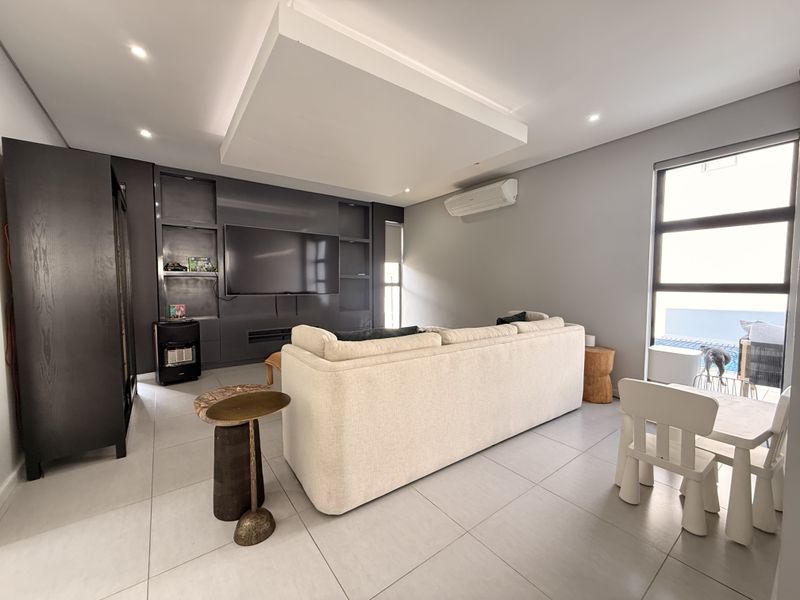 A modern living room featuring a large built-in media unit, recessed ceiling detail, split-system air conditioning, and large windows overlooking the pool area.