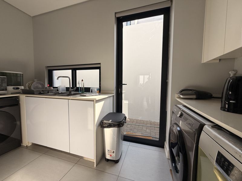 A functional laundry room featuring built-in cabinetry, a stainless steel sink, tiled flooring, and direct access to the exterior via a glass door.