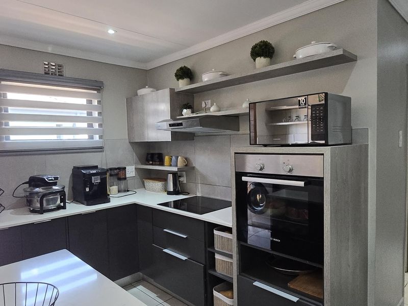 Modern kitchen featuring sleek dark cabinetry, quartz countertops, integrated oven, induction hob, and floating shelving with neutral tiled flooring.