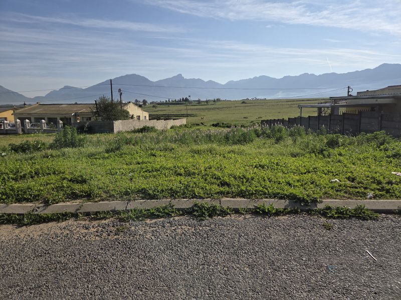 A vacant residential plot featuring a gravel road frontage, concrete boundary walls, and scenic mountain views in the background.
