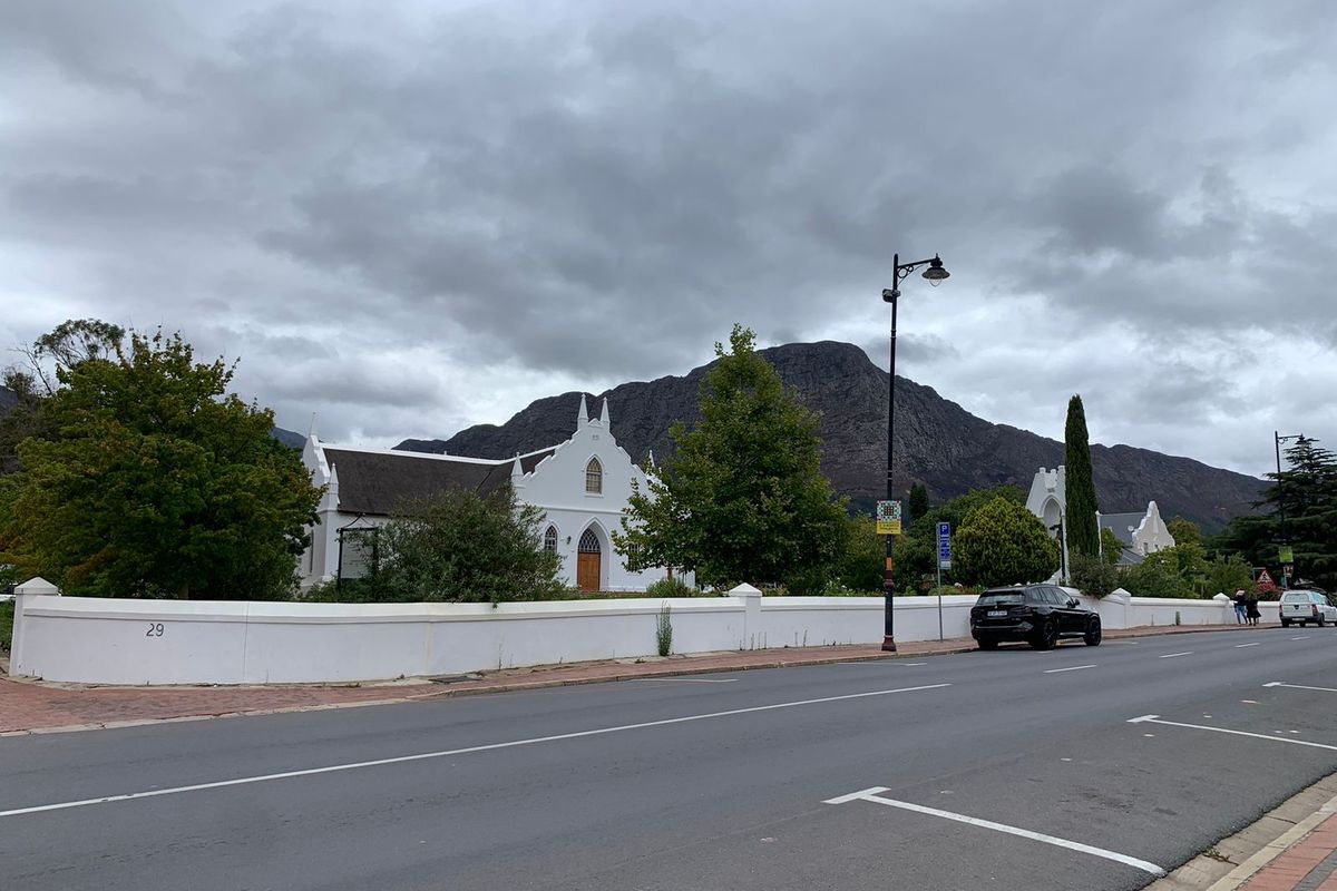 Street view of the property frontage featuring a white boundary wall, mature trees, and mountain backdrop.