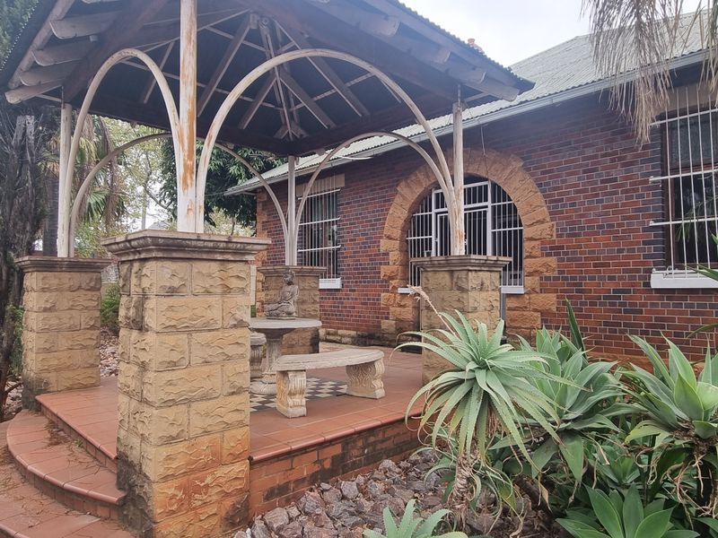 A covered outdoor patio area featuring a brick facade, stone pillars, and a metal gazebo structure, set within a landscaped garden.