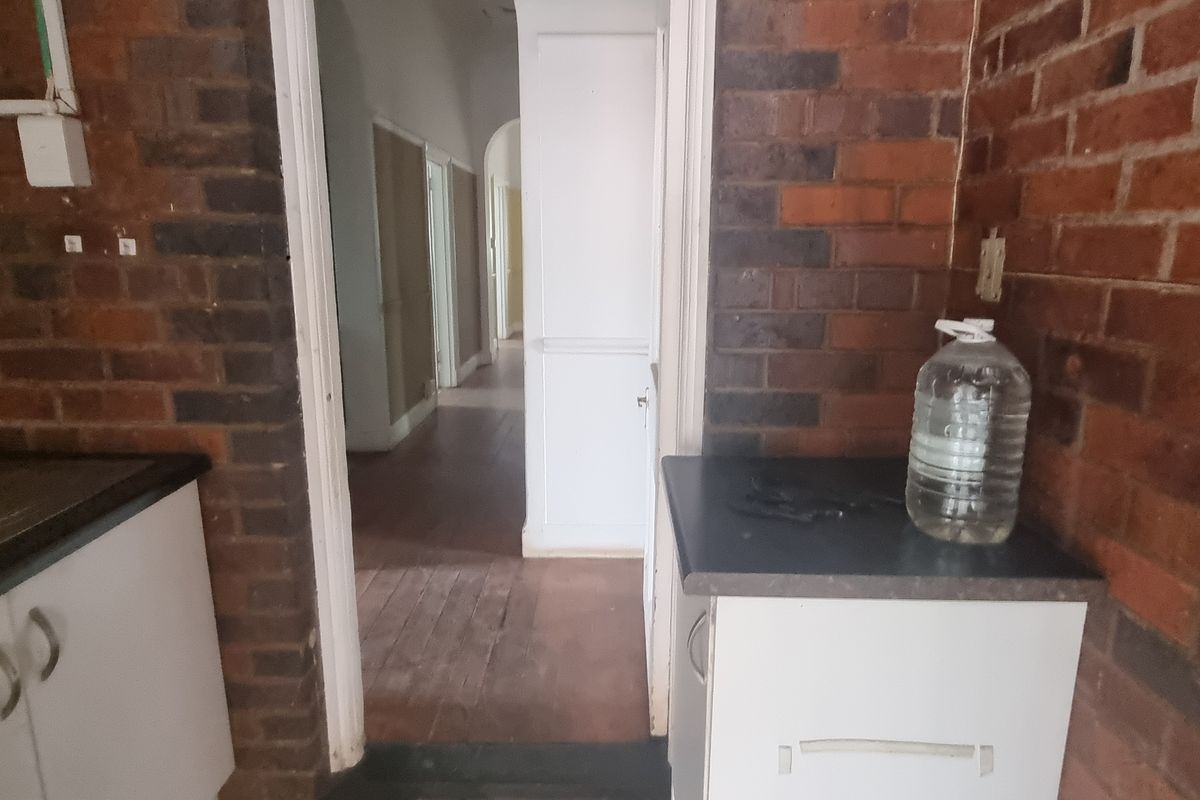 A dated kitchen area featuring exposed brick walls, built-in cabinetry, and timber flooring, leading into a hallway. Requires significant renovation.