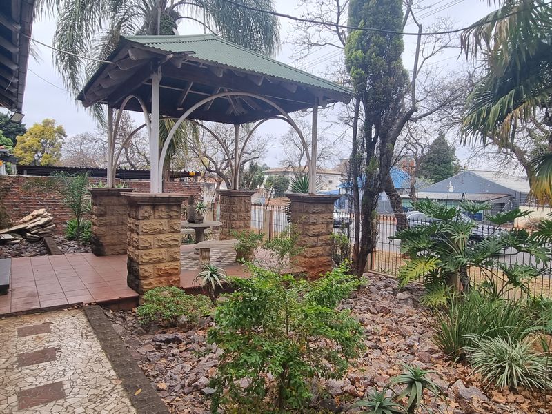 Front garden area featuring a stone gazebo, tiled patio space, and established landscaping adjacent to the brick-clad house exterior.
