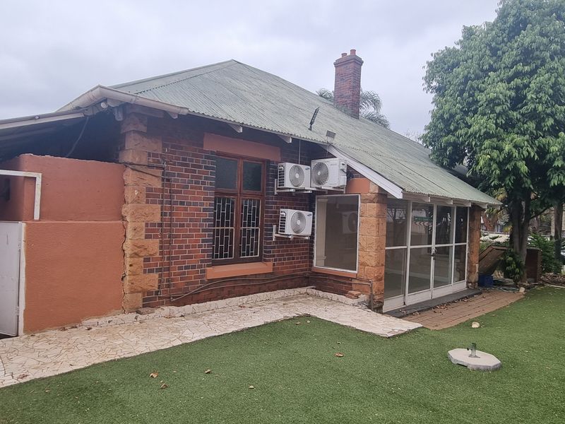 Rear view of a brick house featuring a corrugated metal roof, external air conditioning units, sliding glass doors, and a paved area with artificial lawn.