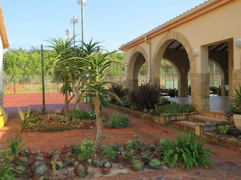 A covered patio area featuring stone-clad arches and brick paving, overlooking a private tennis court with established garden landscaping.