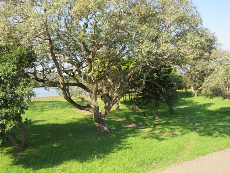 A spacious, well-maintained garden area featuring mature trees, lush green lawn, and a paved pathway leading towards a scenic water view.