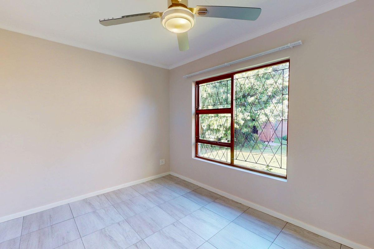 A bright, empty bedroom featuring neutral walls, modern tiled flooring, a ceiling fan, and a large window with security bars.