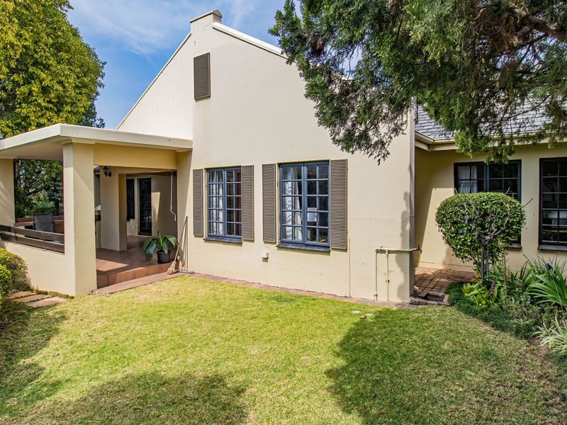 Rear view of the property featuring a covered patio, well-maintained lawn, landscaped garden, and classic casement windows with shutters.