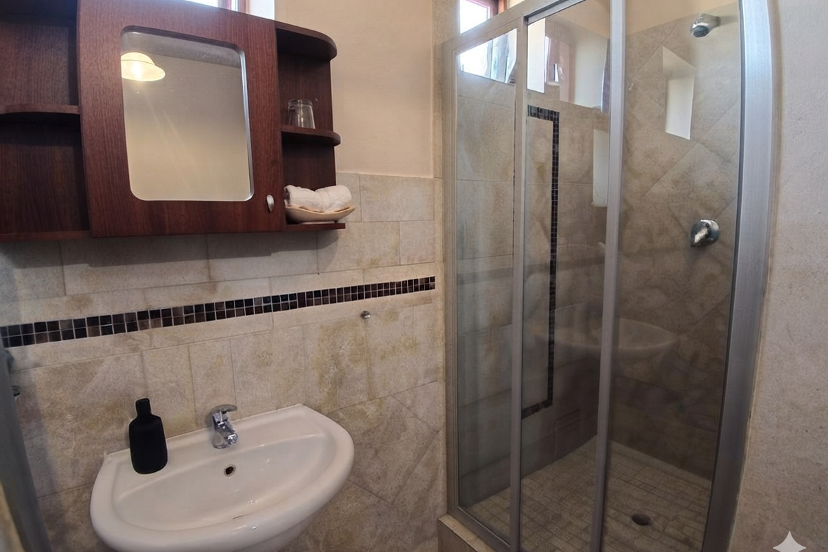 A functional bathroom featuring a pedestal basin, a glass-enclosed shower, and a wall-mounted medicine cabinet with neutral stone-look wall tiles.