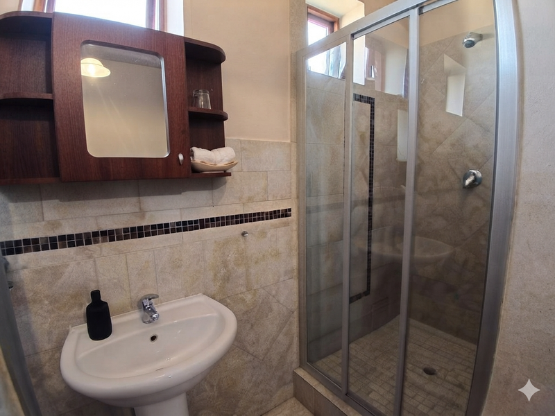 A functional bathroom featuring a pedestal basin, a glass-enclosed shower, and a wall-mounted medicine cabinet with neutral stone-look wall tiles.