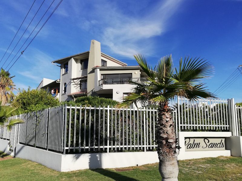 Front elevation of the Palm Sands apartment complex, featuring a secure metal perimeter fence, landscaped garden, and multi-level building structure with balconies.
