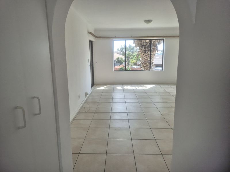 An empty, sunlit living area featuring neutral tiled flooring, an arched entryway, and a large window providing natural light and exterior views.