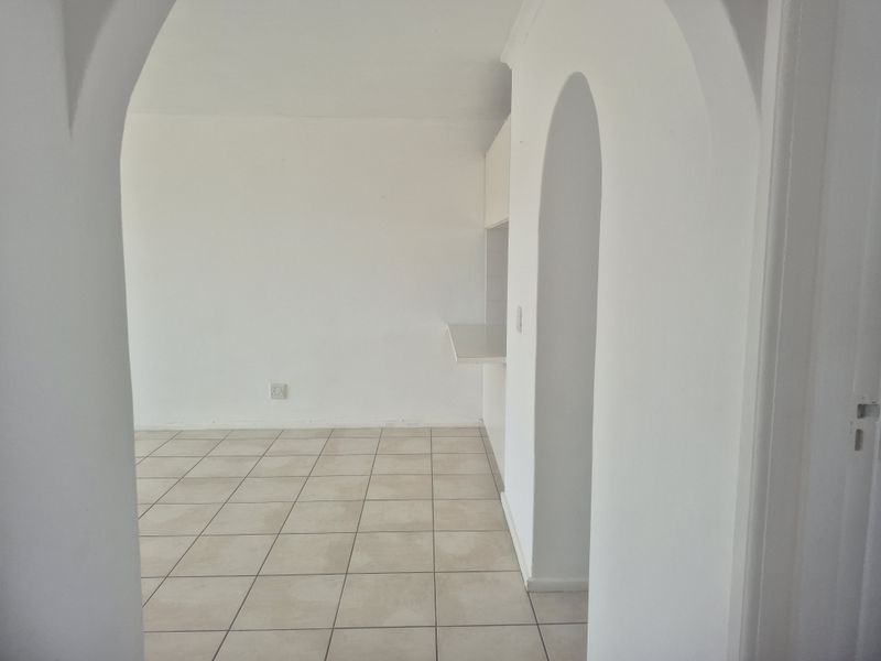 An empty, neutral-toned living area featuring tiled flooring and a distinctive architectural archway leading to a breakfast bar area.