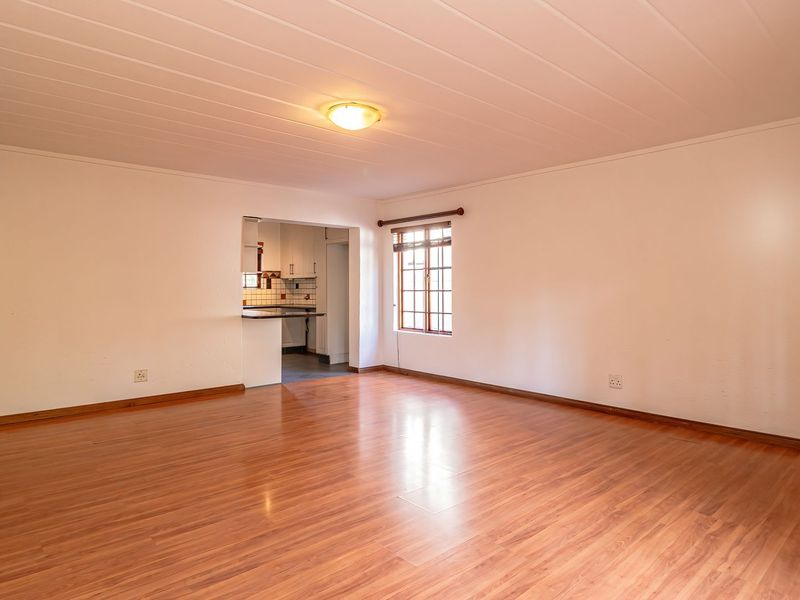 A spacious, empty living room featuring laminate flooring, a timber-framed window, and a neutral interior, with an open-plan view into the adjacent kitchen area.