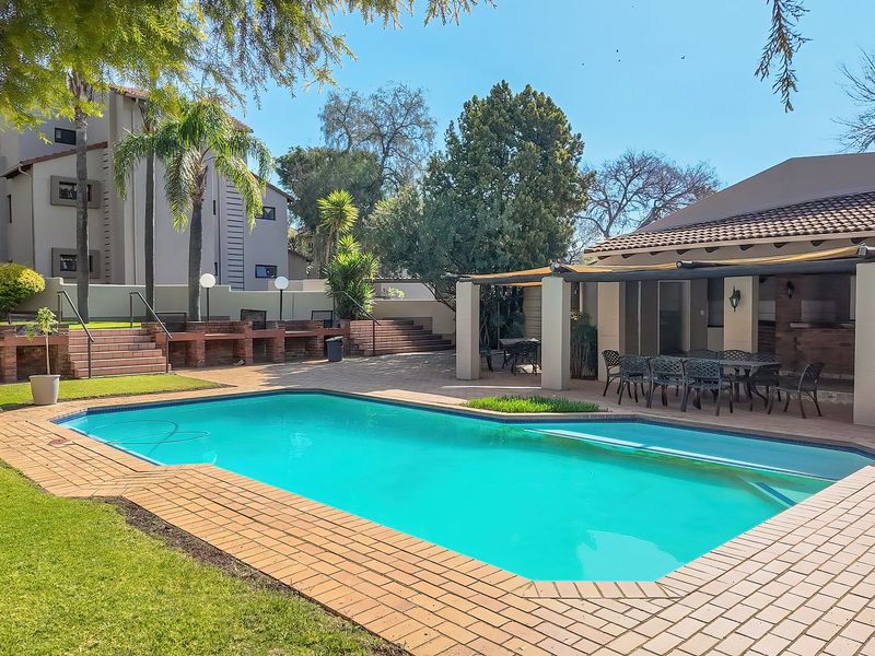 Outdoor pool area featuring a swimming pool, brick paving, and a covered patio with a built-in braai, set within a landscaped garden.