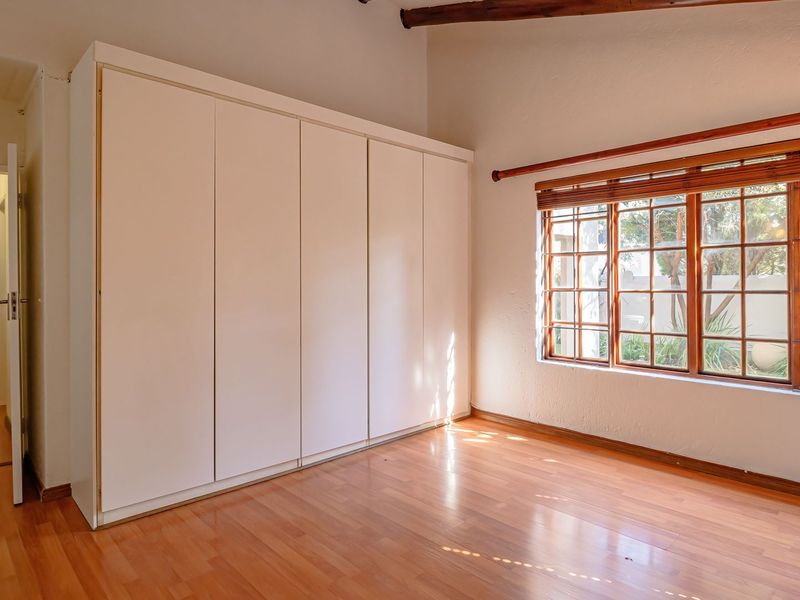A bright, empty bedroom featuring built-in cupboards, warm laminated flooring, and large timber-framed windows with natural light.