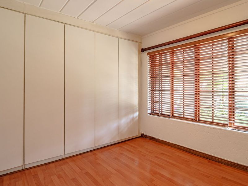 A bright, empty bedroom featuring extensive built-in cupboards, warm laminate flooring, and wooden venetian blinds.