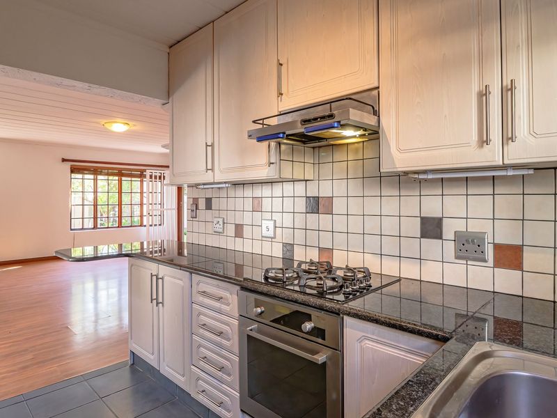 A functional kitchen featuring light wood cabinetry, granite countertops, a gas hob, built-in oven, and a tiled backsplash, flowing into an open-plan living area.