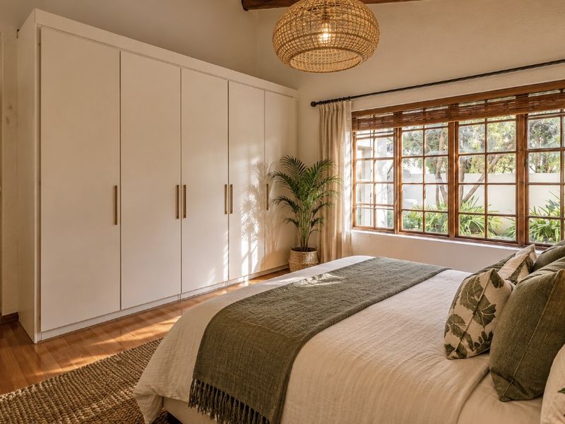 A bright and well-maintained bedroom featuring extensive built-in wardrobes, warm hardwood flooring, and large timber-framed windows overlooking the garden.
