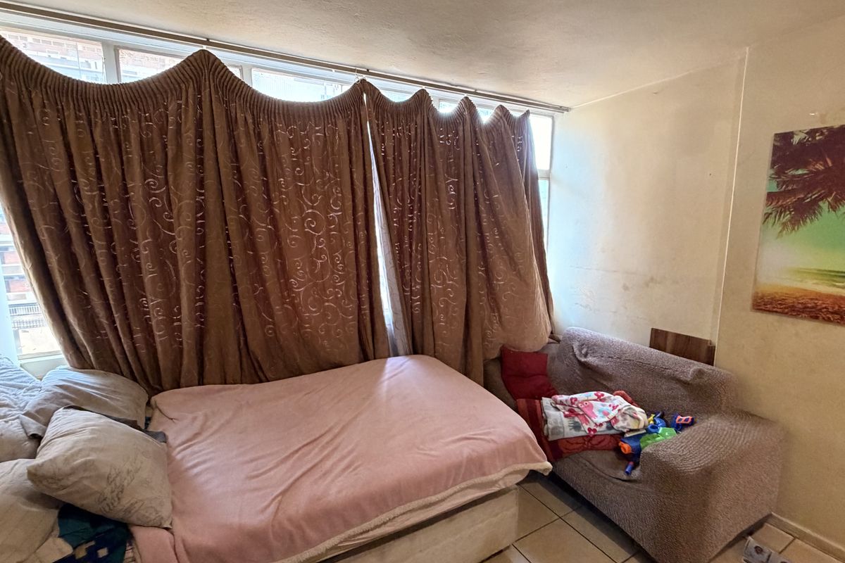 A bedroom featuring tiled flooring and a large window, currently showing signs of wear including wall staining and peeling paint.
