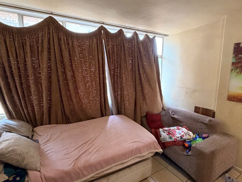 A bedroom featuring tiled flooring and a large window, currently showing signs of wear including wall staining and peeling paint.