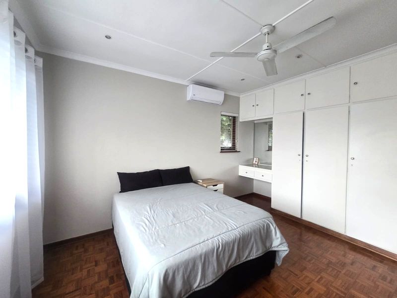 A well-maintained bedroom featuring classic parquet flooring, extensive built-in cabinetry with an integrated vanity, a ceiling fan, and a wall-mounted air conditioning unit.