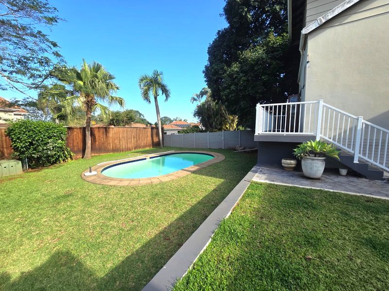A private backyard featuring a swimming pool, a raised deck with white railings, and a well-maintained lawn enclosed by timber and concrete fencing.