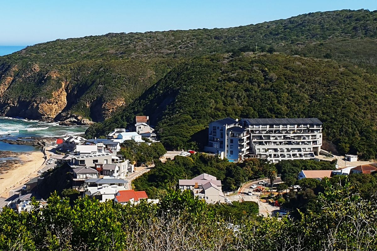 An elevated aerial view of a coastal residential area featuring a large apartment complex nestled against a lush green hillside near the beach.