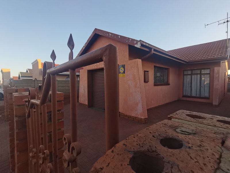 Front exterior view of a single-story house featuring a paved driveway, roller shutter garage door, and secure perimeter fencing.