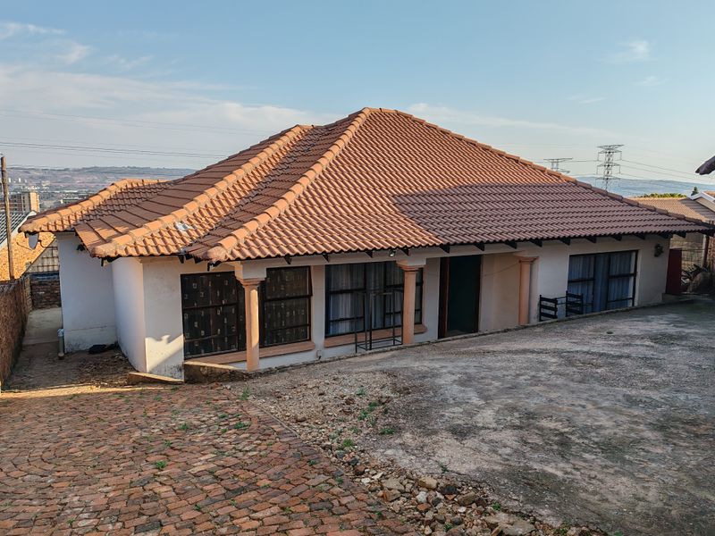 Front elevation of a single-story house featuring a terracotta tiled roof, concrete columns, and a paved driveway area.