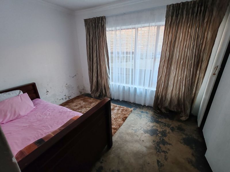 A bedroom featuring a large window and exposed concrete flooring, currently showing significant wear to the wall finishes requiring maintenance and cosmetic updates.