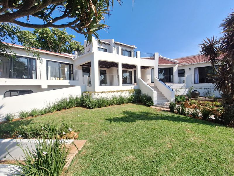 Rear view of a multi-level residence featuring a landscaped garden, covered terrace, upper-level balcony, and white exterior facade with tiled roofing.