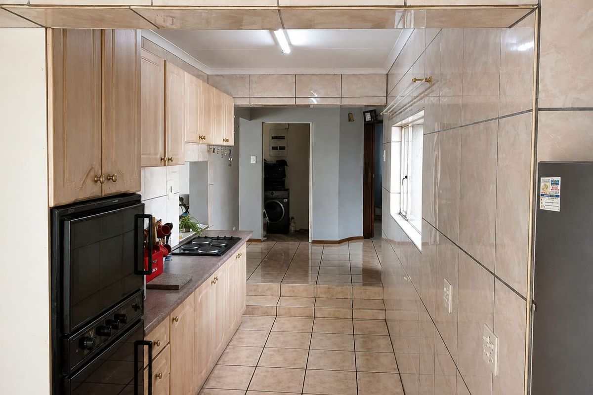 A functional galley-style kitchen featuring light wood cabinetry, built-in oven and hob, and fully tiled walls and floors leading to a utility area.