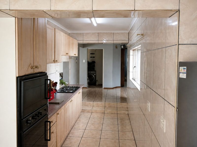 A functional galley-style kitchen featuring light wood cabinetry, built-in oven and hob, and fully tiled walls and floors leading to a utility area.
