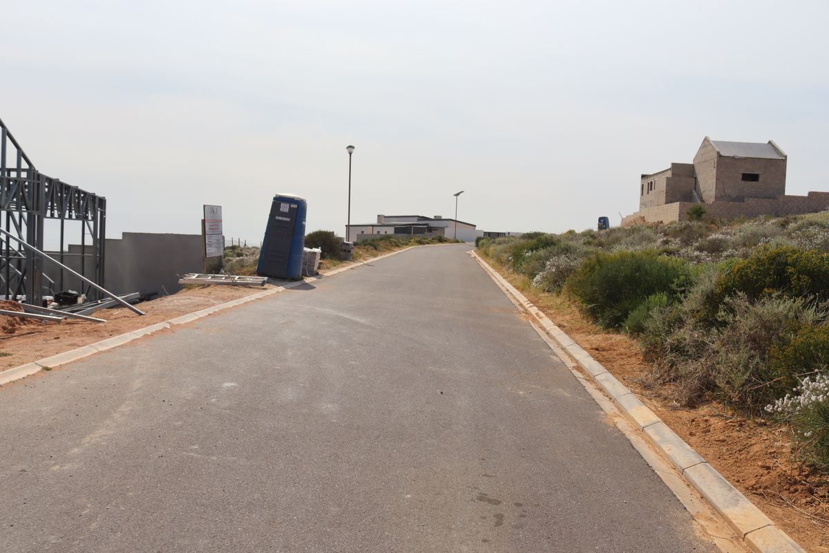 A view of a paved residential street in a developing area, featuring nearby construction framing and surrounding natural vegetation.