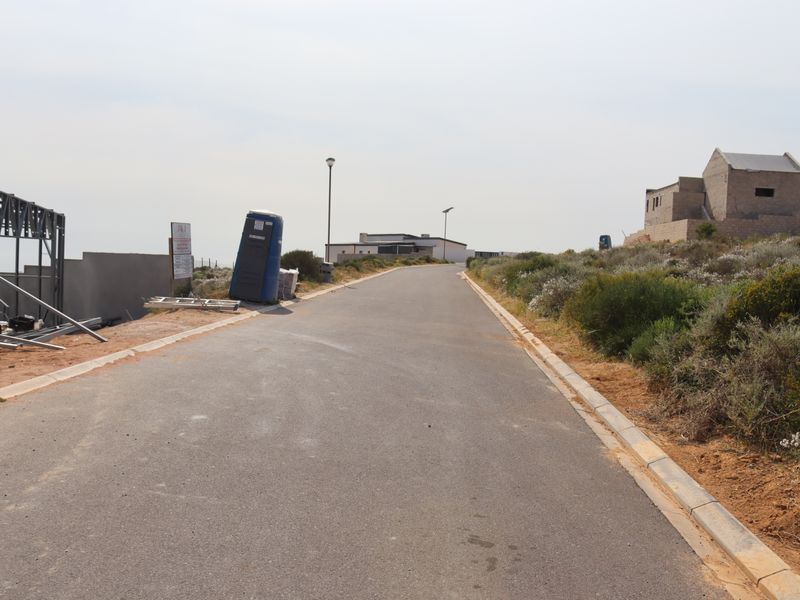A view of a paved residential street in a developing area, featuring nearby construction framing and surrounding natural vegetation.