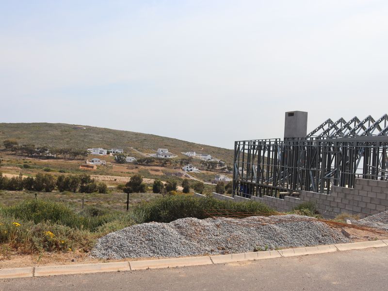 A residential property under construction featuring a steel frame structure and concrete block foundation, situated in a developing landscape.