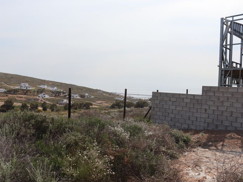 View of a residential plot under construction featuring a steel frame structure, concrete block wall, and wire mesh fencing overlooking the surrounding landscape.