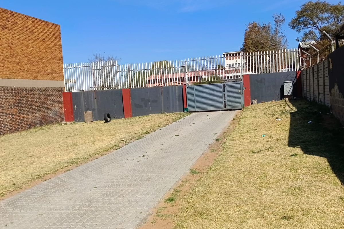 A rear exterior view featuring a paved driveway, perimeter wall with security fencing, and a metal gate.