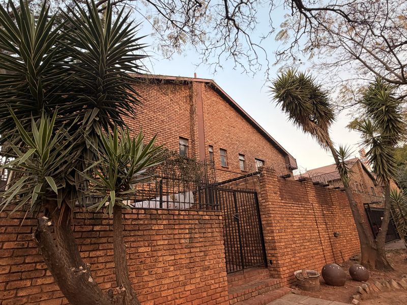 Front exterior view of a townhouse featuring a traditional brick facade, secure perimeter wall with metal gate, and mature landscaping.