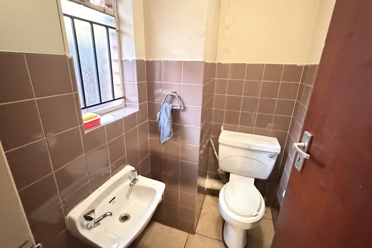 A compact guest bathroom featuring dated ceramic wall tiles, a wall-mounted basin, and a standard toilet with security bars on the window.