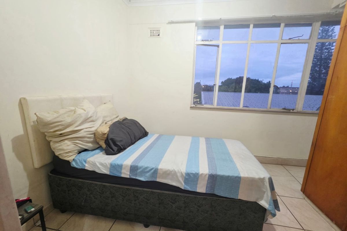 A bedroom featuring tiled flooring, a large multi-pane window, and a built-in cupboard, offering a functional space with potential for cosmetic updates.